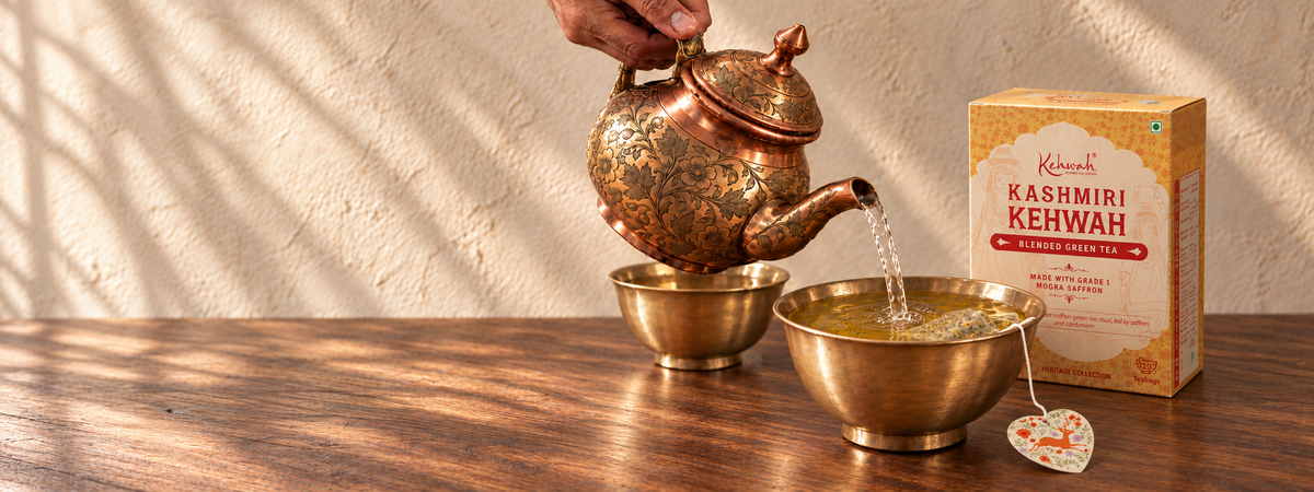 Clear hot water pours from an engraved copper kettle into a brass khosa, with a soft wet tea bag and a Kashmiri Kehwah carton resting behind the cup.