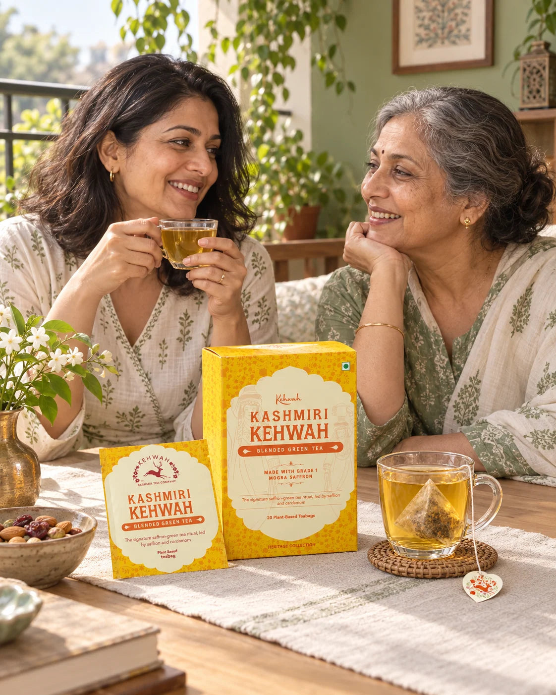 Two women sharing golden Kashmiri Kehwah on a leafy balcony.